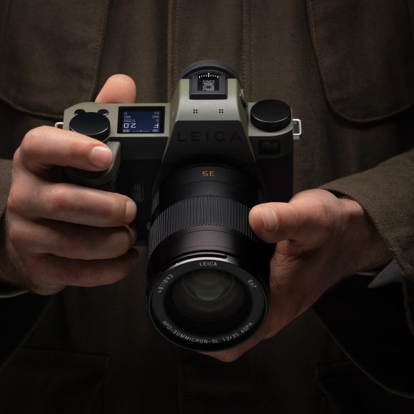 A person in a brown jacket holds a Leica camera with both hands, lens facing forward.