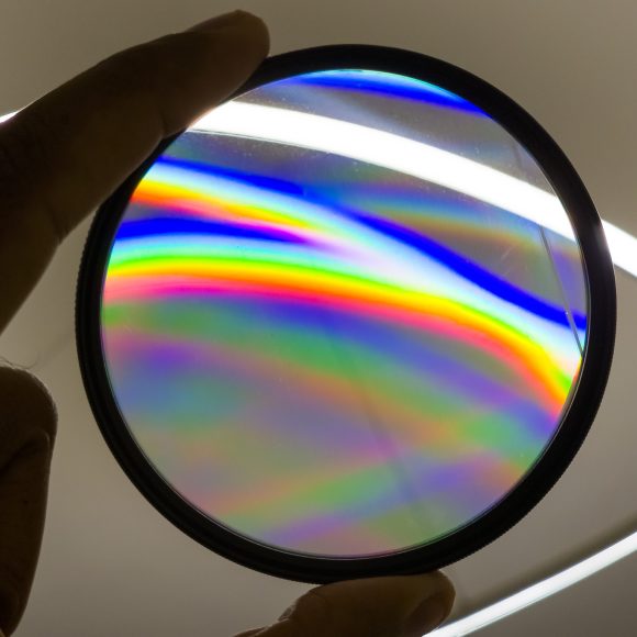 A hand holds a circular glass filter showing rainbow-colored light patterns against a white background.