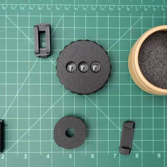 Small black camera lens parts and an open round case on a green cutting mat with a grid pattern.