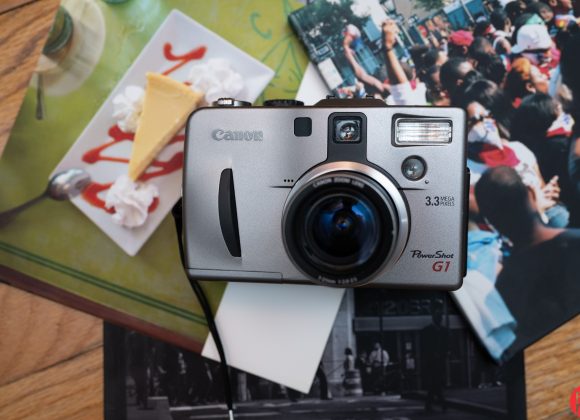 Canon PowerShot G1 camera on magazines with cheesecake and crowd photos on a wooden surface.