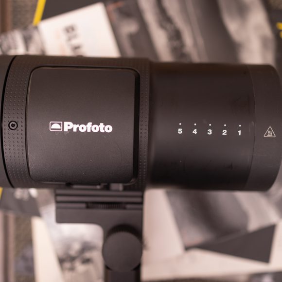 A Profoto studio light is placed on a table with blurred magazines in the background.