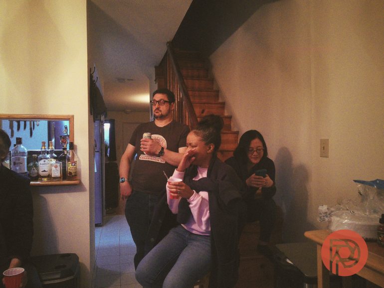 Three people indoors, one standing and two seated, holding drinks near a staircase at a casual gathering.