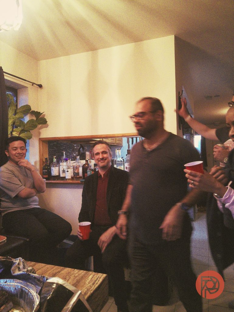 A group of people enjoy drinks and conversation at a cozy indoor gathering near a home bar.