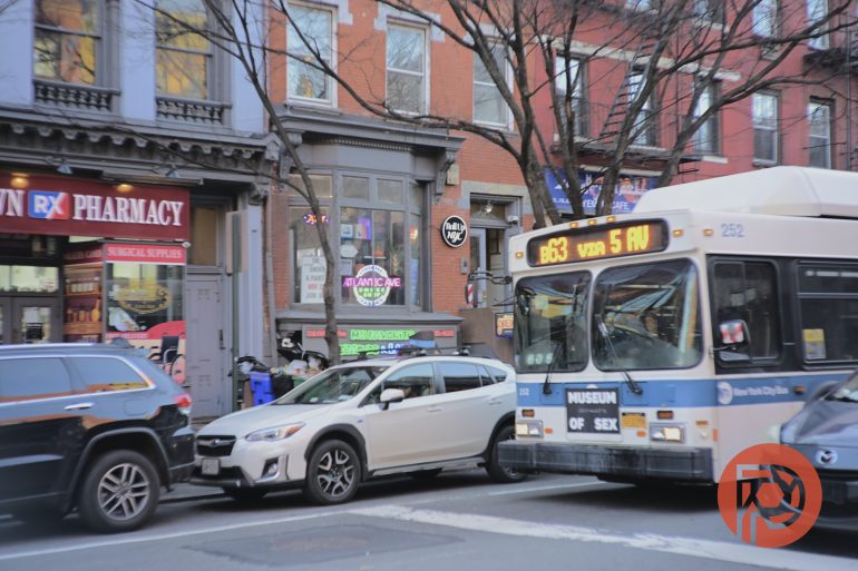 A city street with parked cars, a bus, and storefronts including a pharmacy and the Museum of Sex.