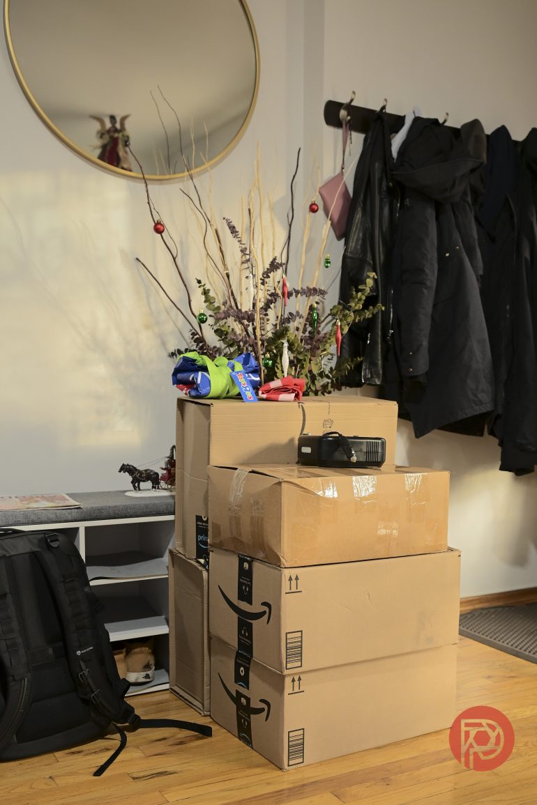 Three stacked cardboard boxes near a coat rack and decorative vase in a sunlit entryway.
