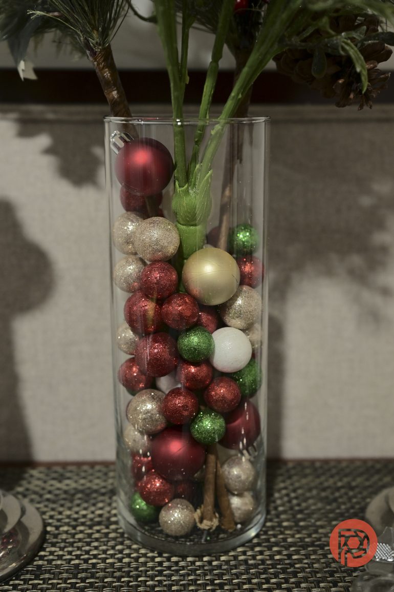 A glass vase filled with festive red, gold, green, and white glittery ornaments and plant stems.