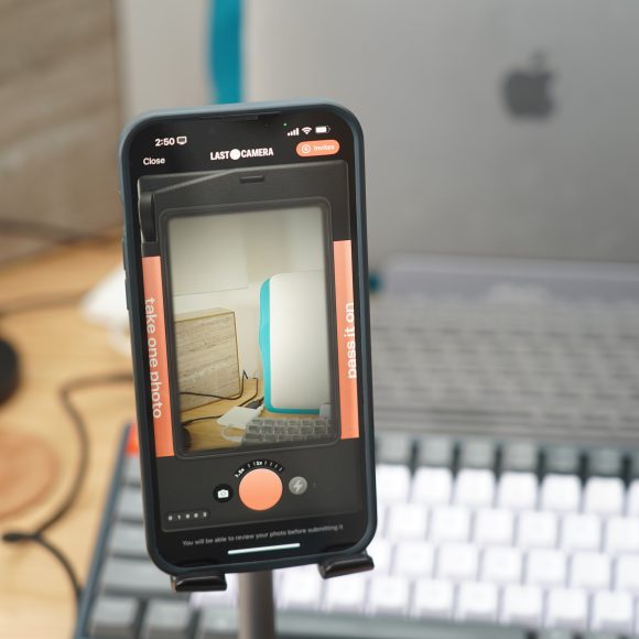 A smartphone on a stand is taking a photo of a laptop and keyboard on a desk.