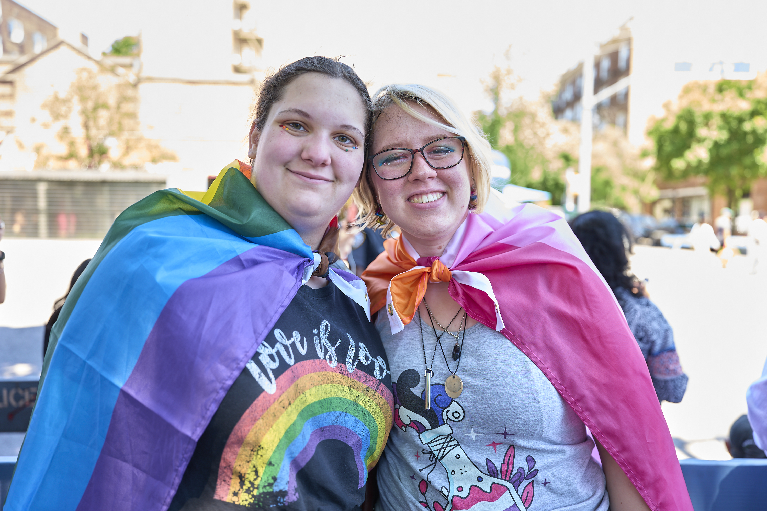Two smiling people in pride capes, posing together outdoors at a daytime LGBTQ+ event.