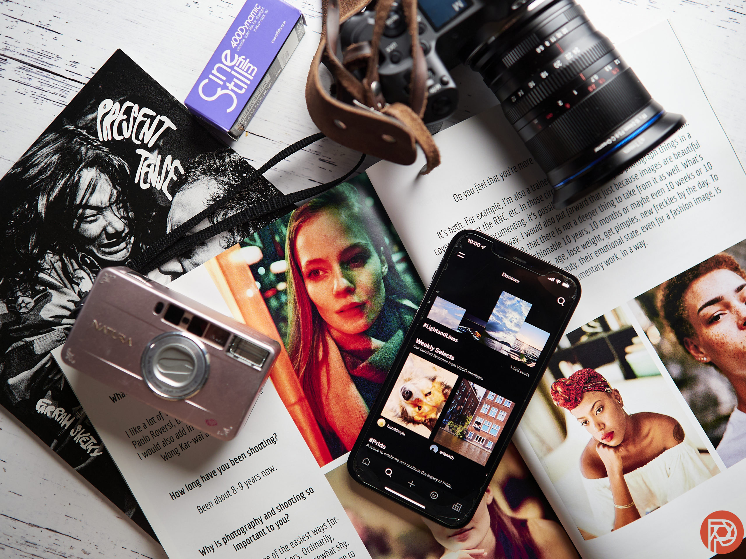 Flat lay of cameras, open magazine, phone with Instagram, lens, and CineStill film on a white surface.