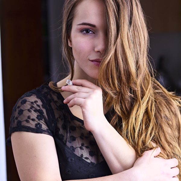 Person with long hair wearing a sheer black top, looking at the camera with one hand near their chin.