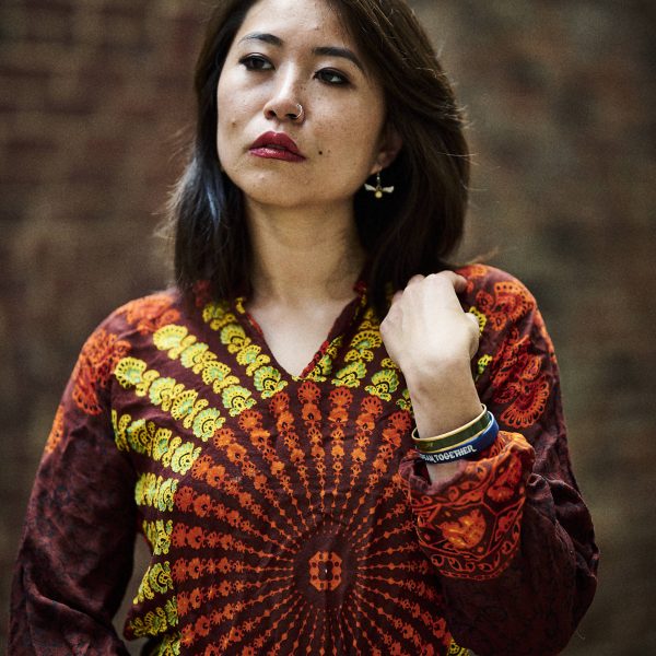 Woman in a vibrant, patterned shirt stands against a brick wall, looking upward thoughtfully.