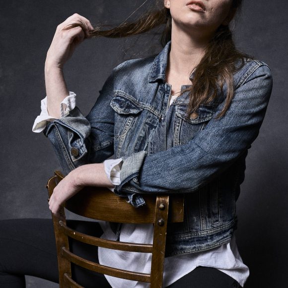 Person in a denim jacket sitting on a wooden chair, holding their hair, with a confident expression.