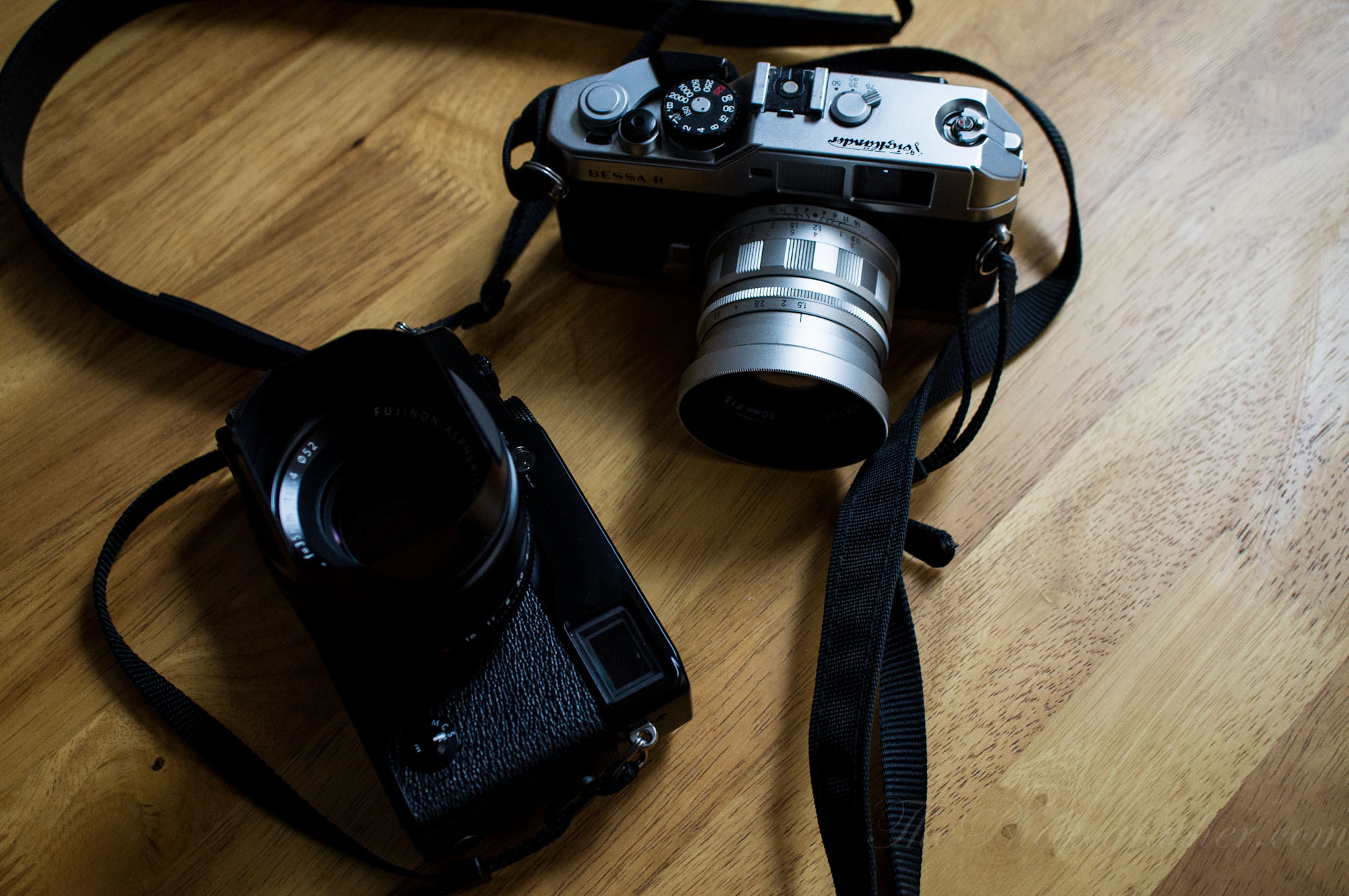Two vintage cameras with straps rest on a wooden table, one silver and one black.