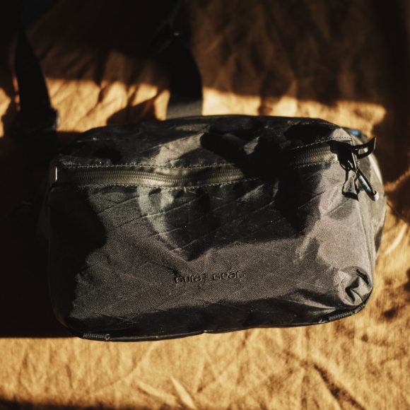 A black sling bag with a front zipper pocket lying on a brown fabric surface in sunlight.