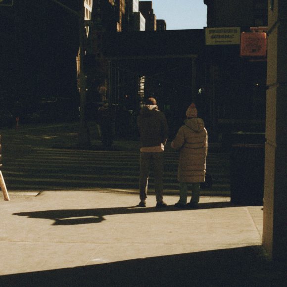 Four people stand and walk on a shadowy city street corner in winter clothing, with sunlight casting long shadows.