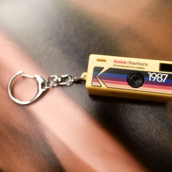 A small Kodak keychain digital camera with 1987 printed on it, lying on a blurred surface.
