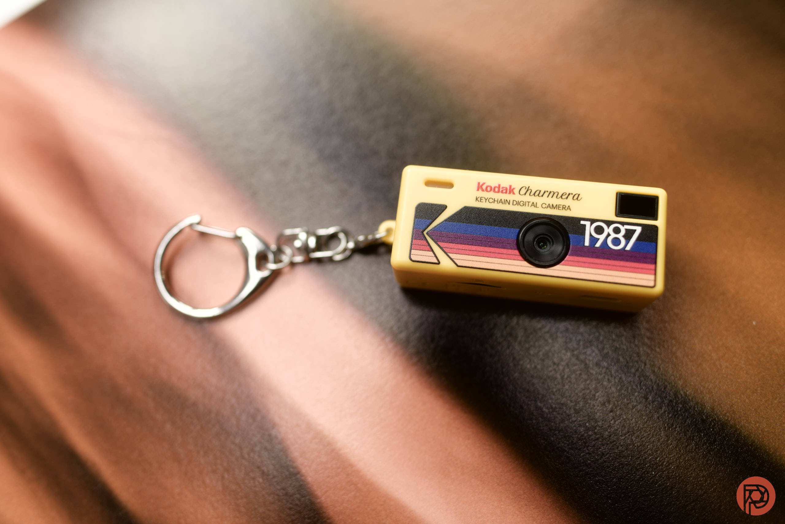 A small Kodak keychain digital camera with 1987 printed on it, lying on a blurred surface.