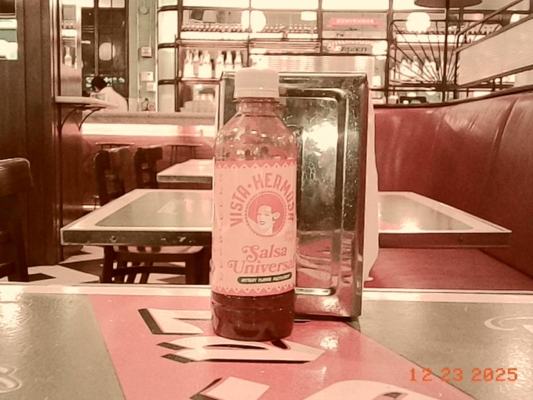 A bottle of salsa sits on a restaurant table with red booths and a napkin dispenser in the background.