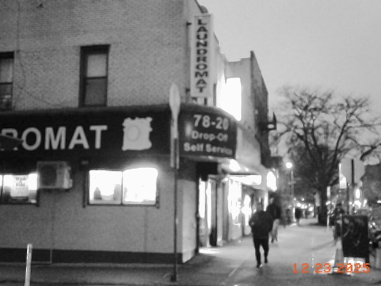 Black-and-white photo of a laundromat on a city street, with people walking on the sidewalk at dusk.