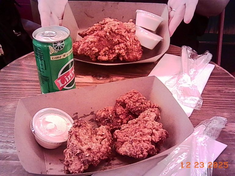 Two trays of fried chicken with dipping sauces, a can of Sprite, and wrapped utensils on a wooden table.