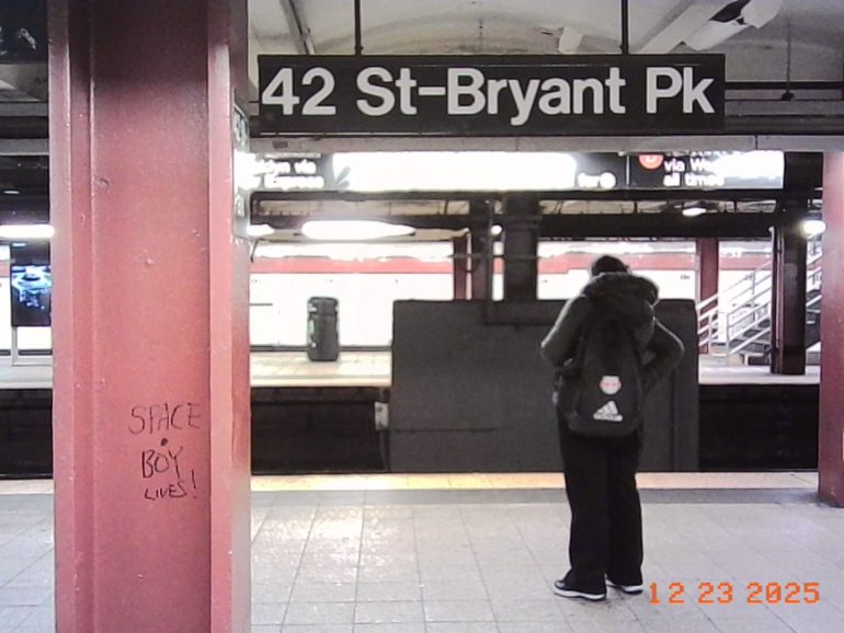 A person stands on a subway platform at 42 St-Bryant Park; graffiti reads SPACE BOY LIVES! on a pillar.