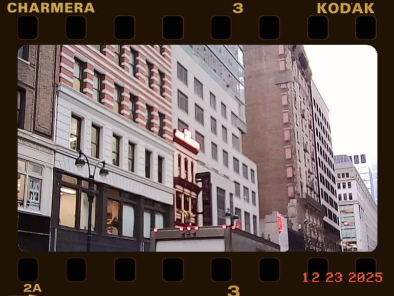 City street with tall buildings, neon signs, and a film frame border marked “KODAK” and dated “12 23 2025.”.