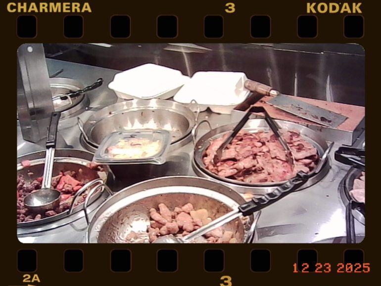 Buffet with metal trays of assorted cooked meats and tongs, plus two white takeout containers in the background.