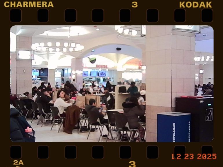 A busy food court with people dining at tables under bright lights, film frame overlay and date stamp.
