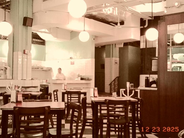 A nearly empty café with black chairs, round hanging lights, and one person behind the counter.