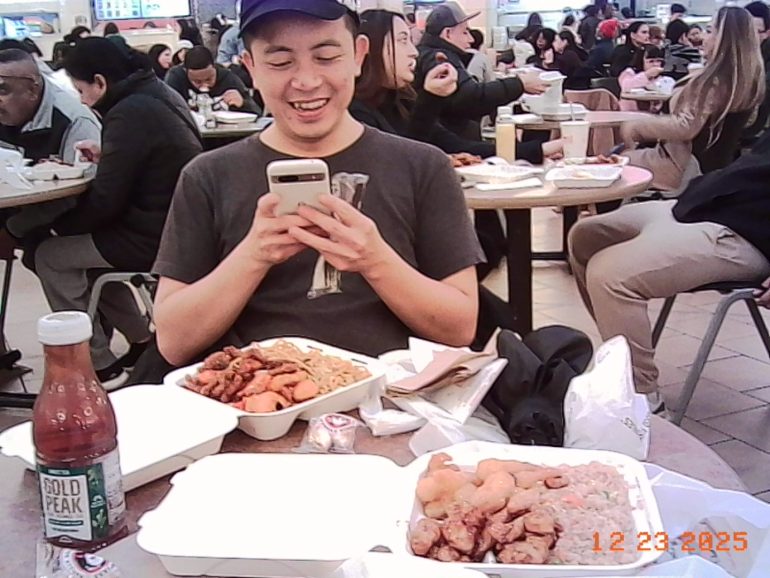 A man smiles at his phone while sitting at a food court table with takeout food and drinks.