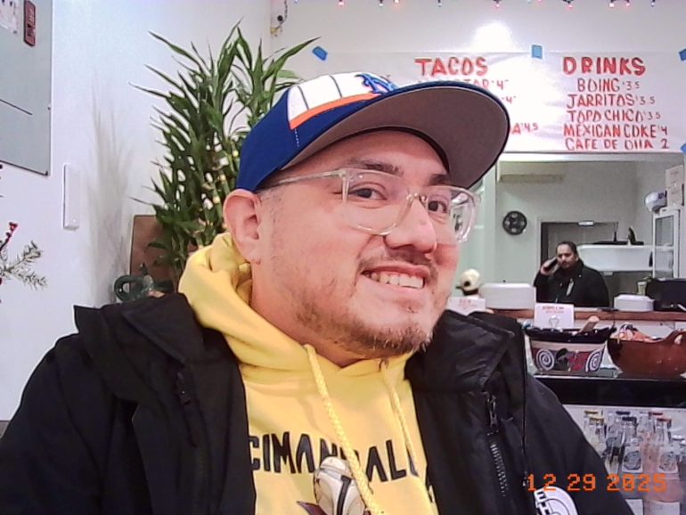 A smiling man in glasses and a cap sits in a restaurant with a menu on the wall behind him.