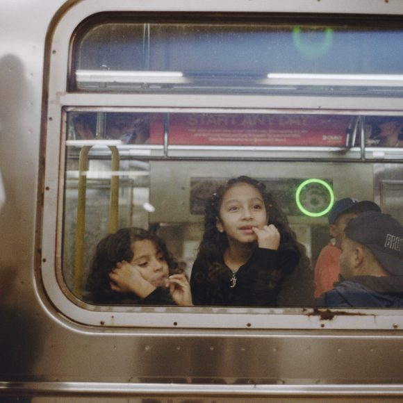 Three children look out the window of a subway train, one girl smiling and touching her chin.