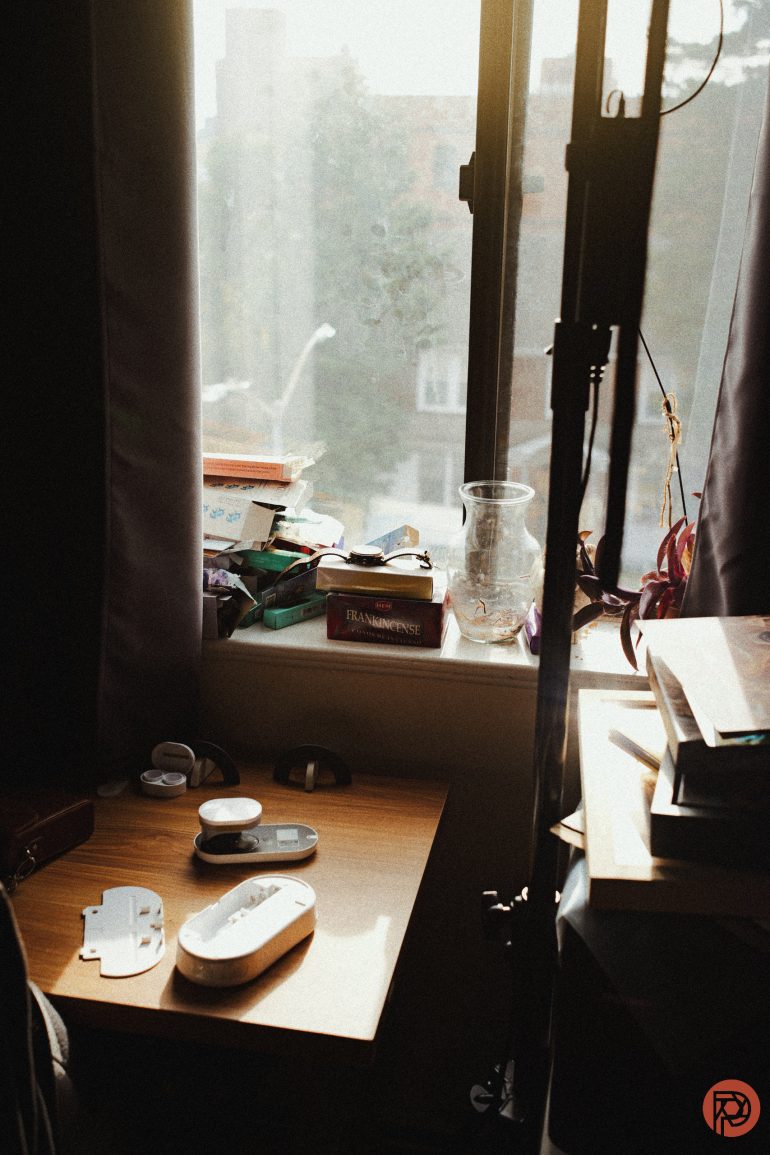 Sunlit cluttered desk by a window with books, electronics, glass, and scattered items in warm light.