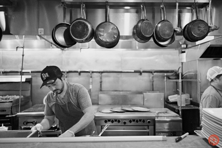 A man in a kitchen prepares food, with pans hanging above a stove and another person in the background.
