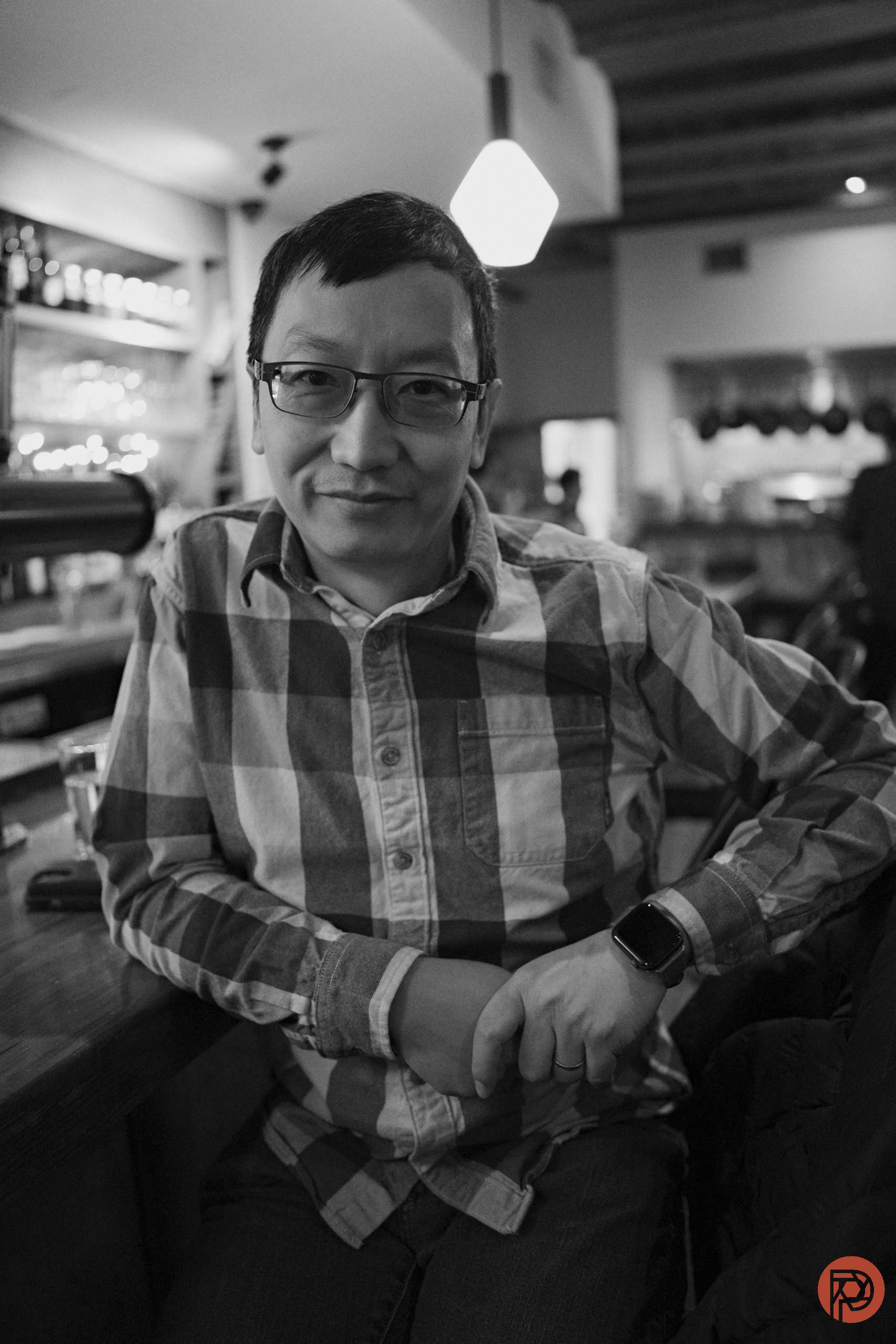 A person in glasses sits at a bar, smiling, in a black and white photo with a relaxed atmosphere.