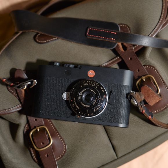 Black Leica camera resting on a khaki and brown leather strap bag, photographed from above.