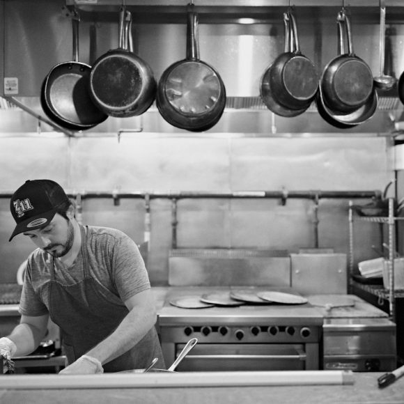 A man cooks in a commercial kitchen with hanging pans and an industrial stove in the background.