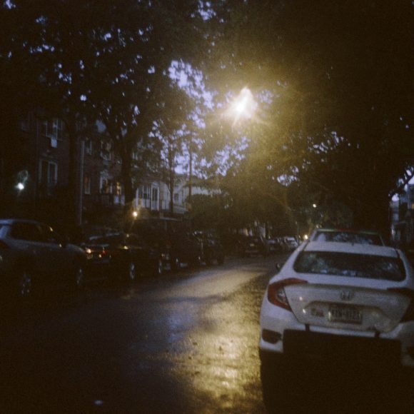 A dimly lit street at dusk, with parked cars and wet pavement reflecting a streetlamp’s glow.