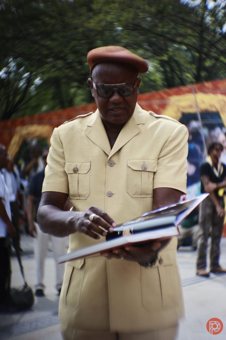 A man in a tan outfit and beret signs a book outdoors, surrounded by people and trees.