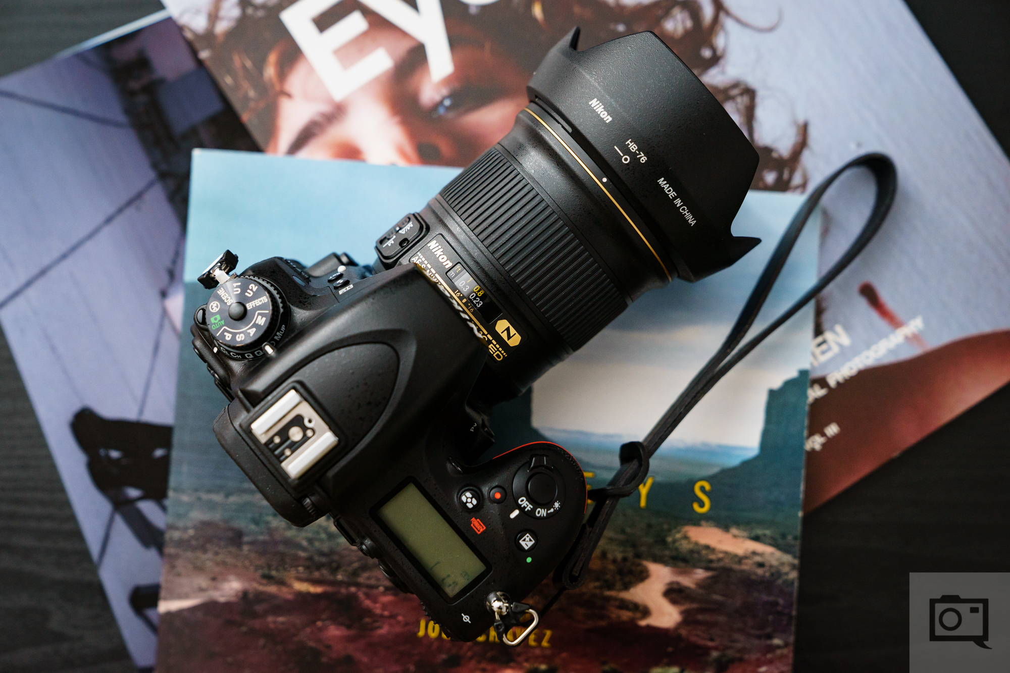 A Nikon DSLR camera with a zoom lens rests on top of colorful photography magazines on a dark surface.