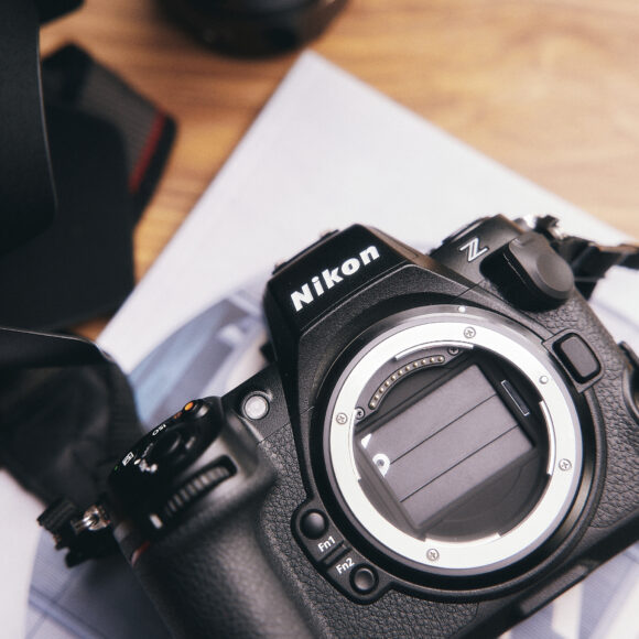 A Nikon Z camera body without a lens sits on a desk, next to a lens hood and some papers.
