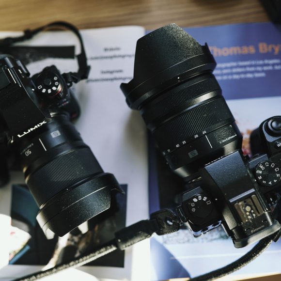 Two black Lumix cameras with large lenses rest on open books and magazines on a wooden surface.