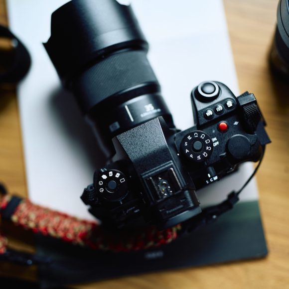 A black digital camera with a large lens sits on a wooden table next to a camera strap and another lens.