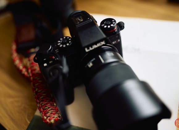 Close-up of a black Lumix camera with a lens, placed on a wooden surface next to a braided camera strap.