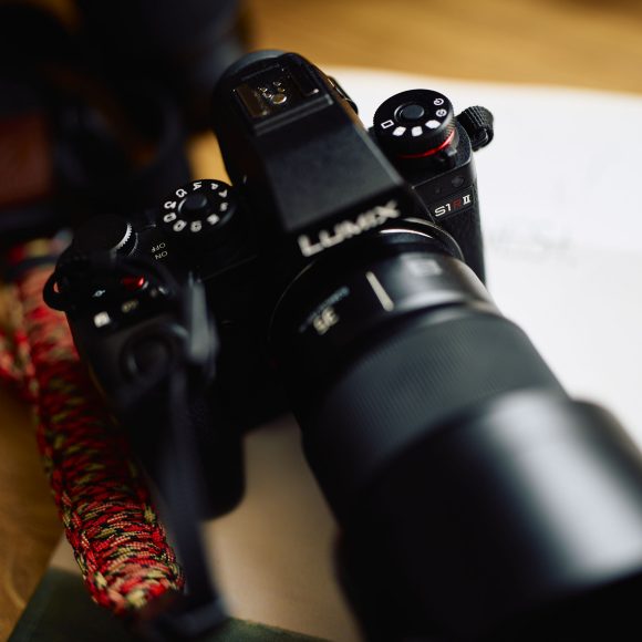 Close-up of a black Lumix camera with a lens, placed on a wooden surface next to a braided camera strap.