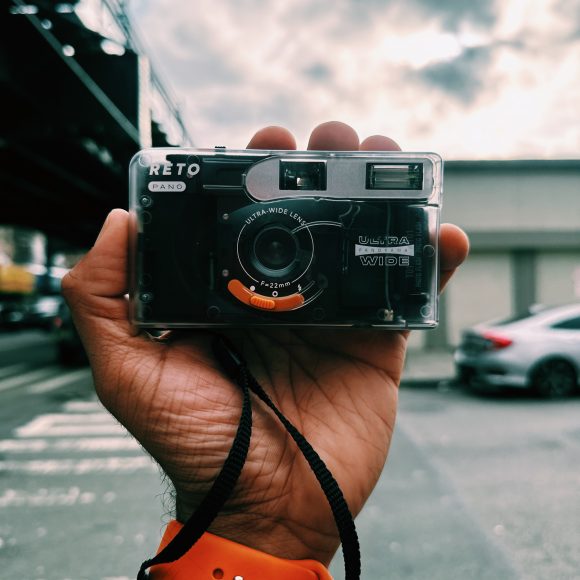 A hand holding a retro film camera outdoors, with cars and cloudy sky in the background.