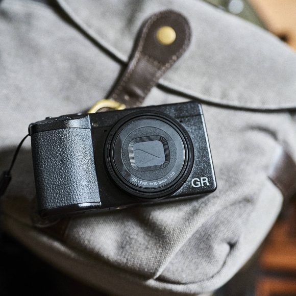A compact black digital camera resting on a light gray canvas bag with leather details.