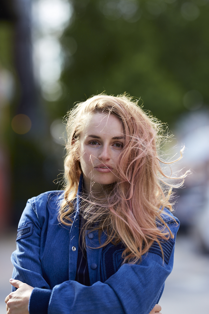 Woman with long blonde hair stands outdoors in a blue jacket, with wind blowing her hair across her face.