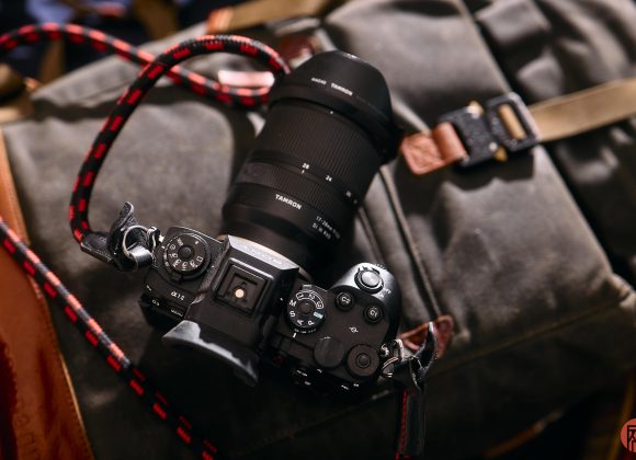 A digital camera with a large lens rests on a gray backpack with brown leather straps.