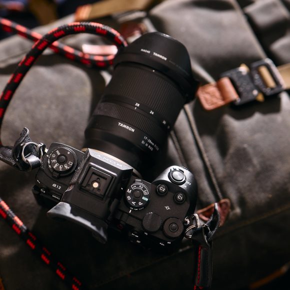 A digital camera with a large lens rests on a gray backpack with brown leather straps.